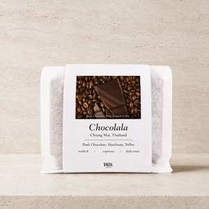 a bag of thai coffee beans in a translucent bag with a white paper sleeve with the words chocolala on it on a stone background and ledge made for espresso