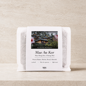 a bag of thai coffee beans in a translucent bag with a white paper sleeve with the words mae au kor on it on a stone background and ledge made for filtered and espresso coffee