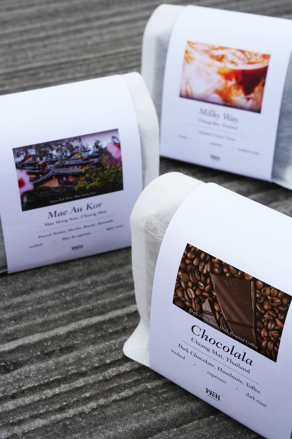 3 bag of thai coffee beans in a translucent bag with a white paper sleeve with the words Mae Au Kor, Milky Way, and Chocolala on it on a grey concrete floor