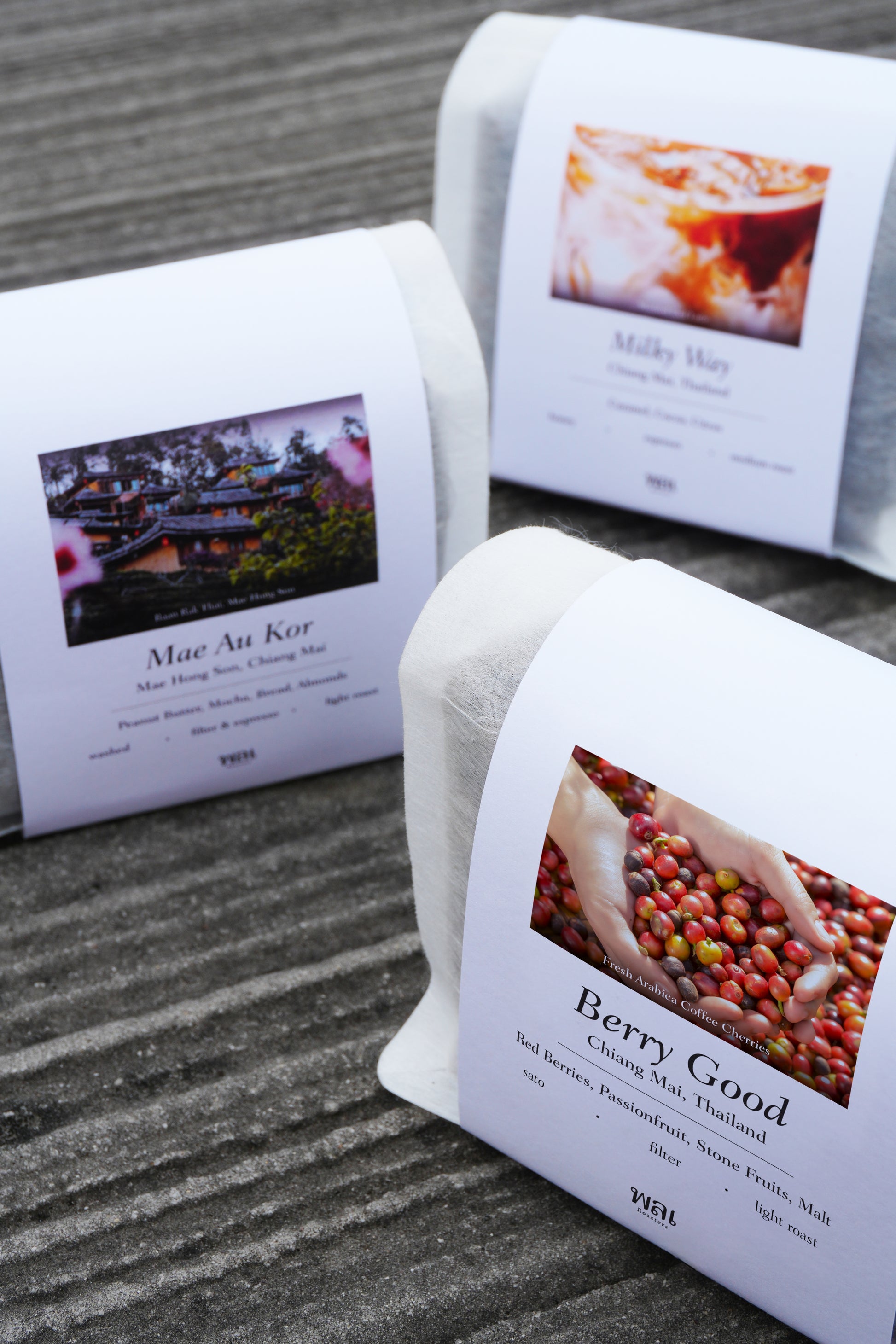 3 bag of thai coffee beans in a translucent bag with a white paper sleeve with the words Mae Au Kor, Milky Way, and Berry Good on it on a grey concrete floor
