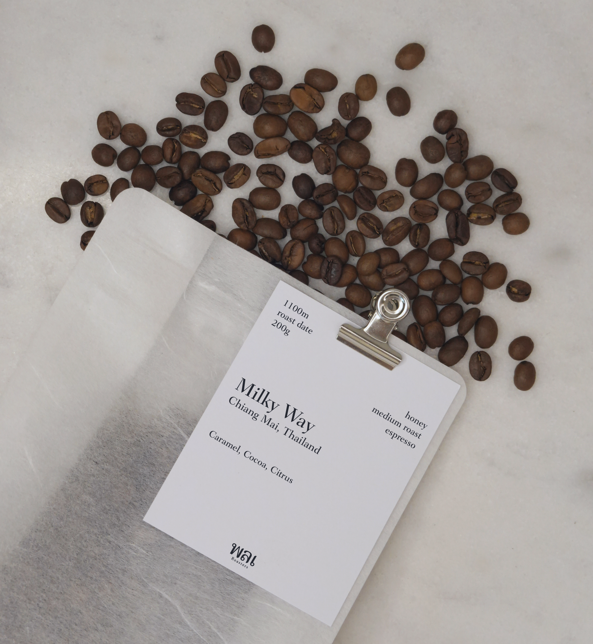 transparent/translucent bag of medium roast thai coffee beans from wai roasters with a card/postcard/polaroid clipped onto the bag roasted for subscriptions and coffee beans spilled on marble table
