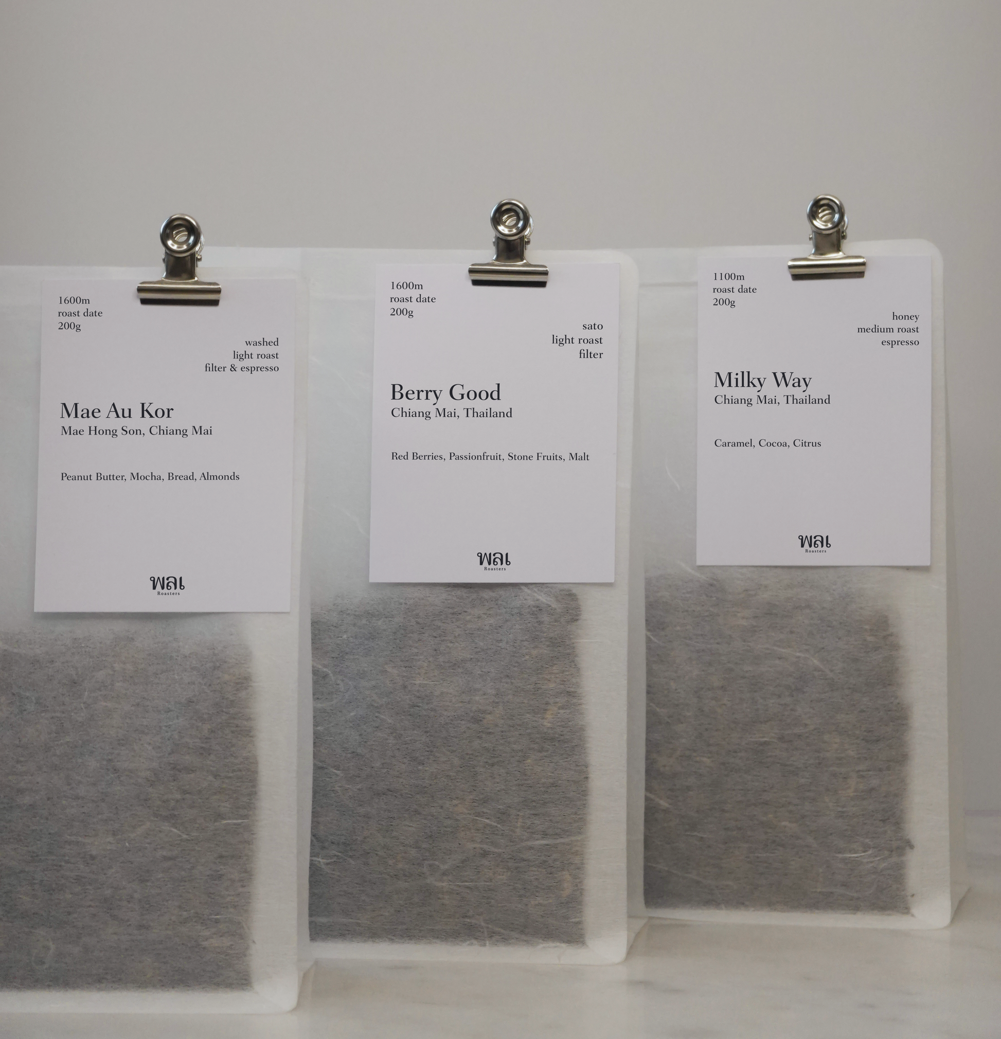 three transparent/translucent bags of light/medium roast thai coffee beans from wai roasters with a card/postcard/polaroid clipped onto the bag roasted for subscriptions
