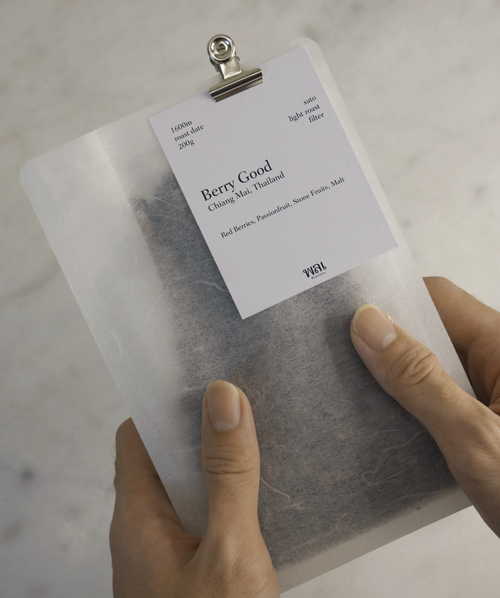 transparent/translucent bag of light roast thai coffee beans from wai roasters with a card/postcard/polaroid clipped onto the bag roasted for subscriptions being held in hands on top of marble
