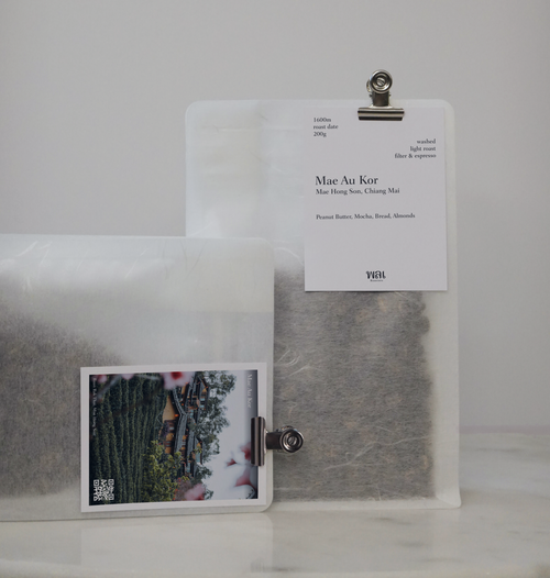 A transparent/translucent bag of light roast thai coffee beans from wai roasters with a card/postcard/polaroid clipped onto the bag roasted for filter brews