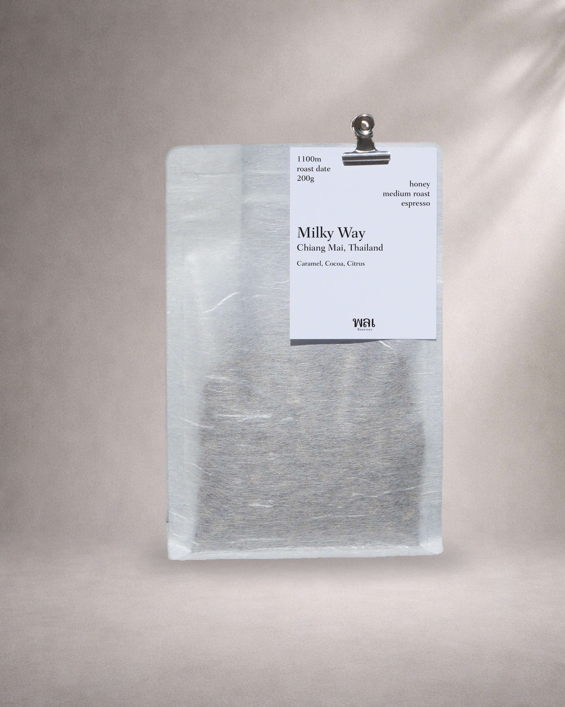 a bag of thai coffee from thailand in a translucent/transparent bag with llinen like texture, withy a meta bulldog cllip holding a card on the bag with a grey background roasted for espresso coffee