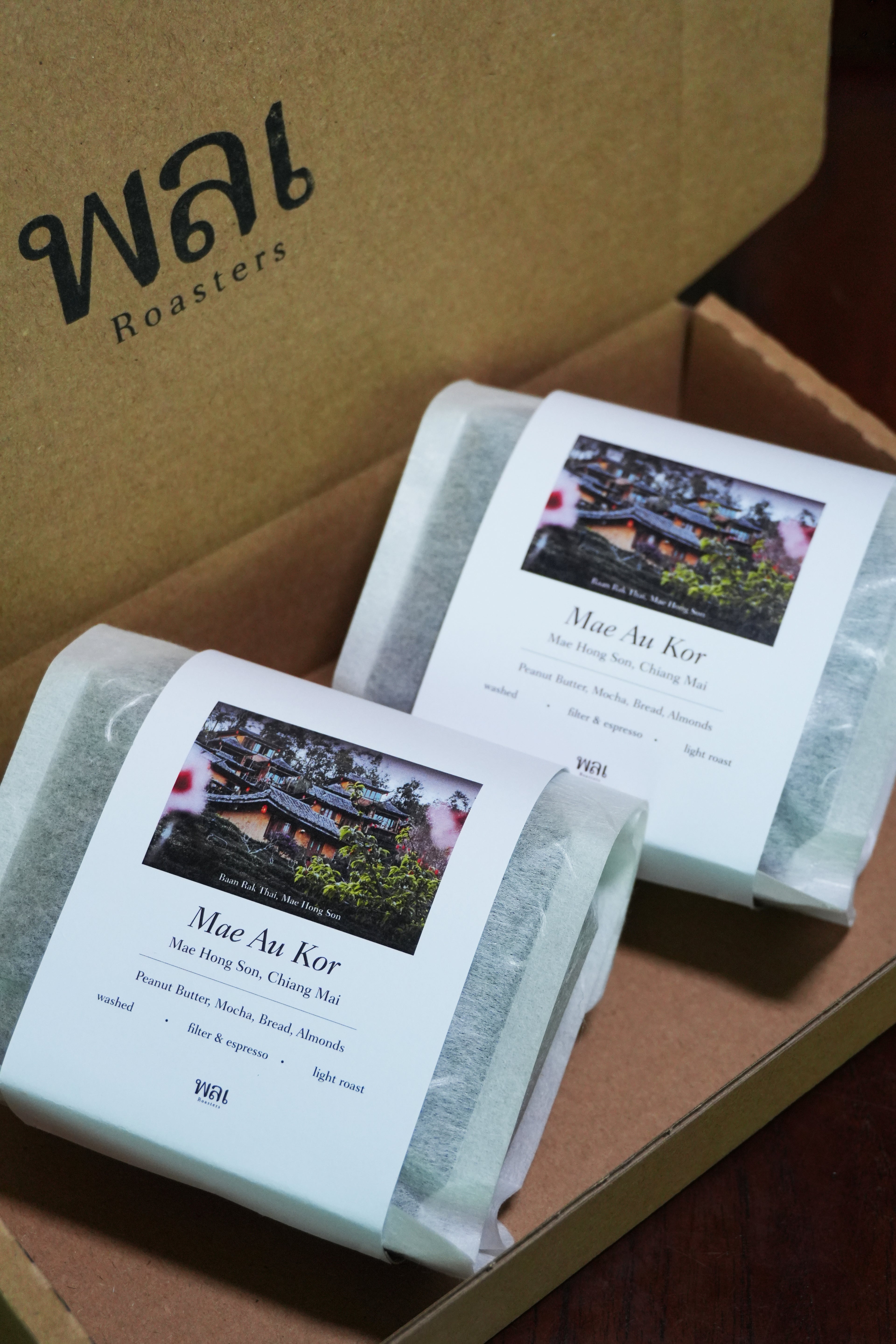 2 bag of thai coffee beans in a translucent bag with a white paper sleeve with the words Mae Au Kor, on it on in a pizza style cardboard box with black logo stamped on for wai roasters