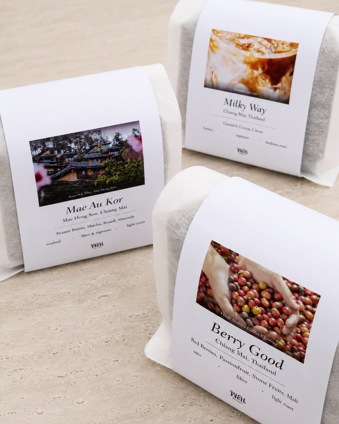 3 bag of thai coffee beans in a translucent bag with a white paper sleeve with the words Mae Au Kor, Milky Way, and Berry Good on it on a grey concrete floor